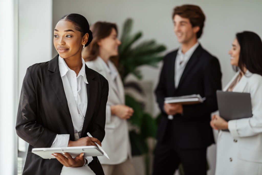A business professional looking positively outside her office window, with her colleagues talking in the background.